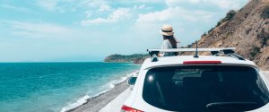 Student traveller in a hat looking out to sea from a parked car on a coastal road – ideal for a budget road trip.
