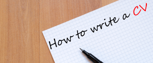 Hand-written note saying 'How to write a CV' on grid paper with black pen on wooden desk
