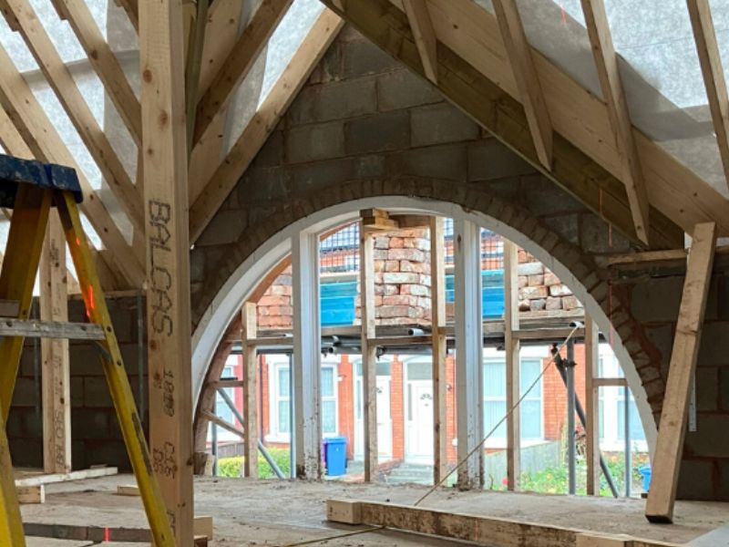 Interior of a Liverpool property mid-restoration, showing a newly constructed timber roof structure and insulation above a preserved original curved brick archway, with a worker on a stepladder to the left.