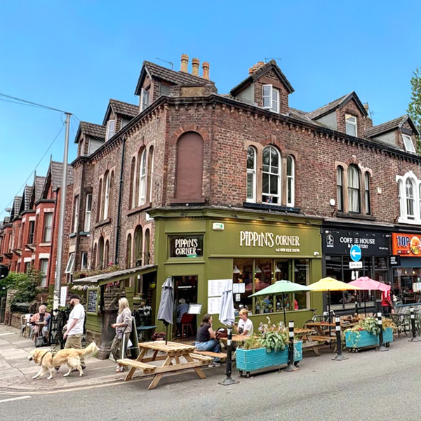 Sunny street corner on Lark Lane in Aigburth, with a green-fronted café, outdoor tables under colourful parasols, and people relaxing along the pavement.
